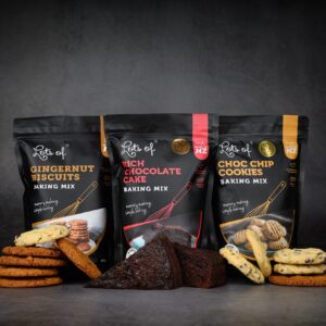 Selection of Lots of Baking Mixes products including Gingernut Biscuits, Rich Chocolate Cake, and Choc Chip Cookies baking mixes with freshly baked treats displayed in front on a dark background.